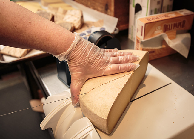 About Us.Kristi cutting cheese.650x464.72res This is a picture of Fromagination Cheesemonger Kristi cutting cheese behind the counter in the shop.