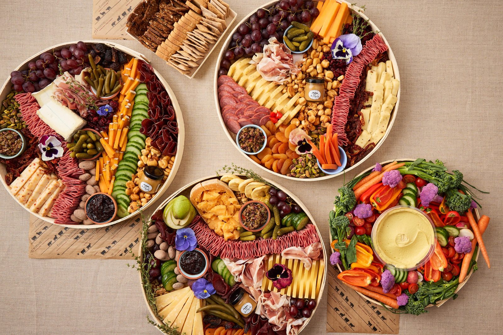 corporate-catering-cheeseboards-veggie-tray-closer Four round catering platters of cheese, charcuterie, fruit, and vegetables with dip.