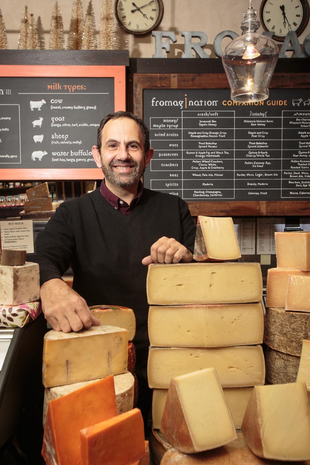 our-story-ken-monteleone-full-frame Fromagination owner Ken Monteleone behind a counter stacked with wheels of cheese.